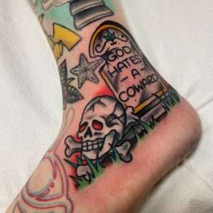 A custom traditional tattoo on a heel, with a tombstone that reads "God Hates a Coward," flanked by a skull and crossbones, and tied together with grass underneath, a custom piece by Jacksonville tattoo artist Tim Shafer.