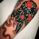 An American traditional Monmon cat tattoo, a black cat tattooed with red flowers and green leaves, holding a snake in its mouth, by artist Zach Westcott in Jacksonville.