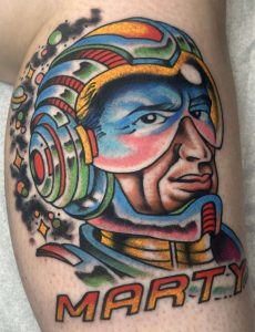 A new school or neo-traditional tattoo of a man in a space helmet with the custom lettering "MARTY" underneath, with a dark space theme background of planets and stars, a custom piece by Jacksonville tattoo artist Ivy Tôn.